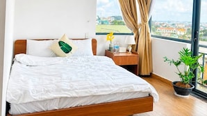 Habitación de hotel moderna con una cama grande, una mesita de noche, una planta en maceta y vistas de la ciudad a través de un ventanal amplio.
