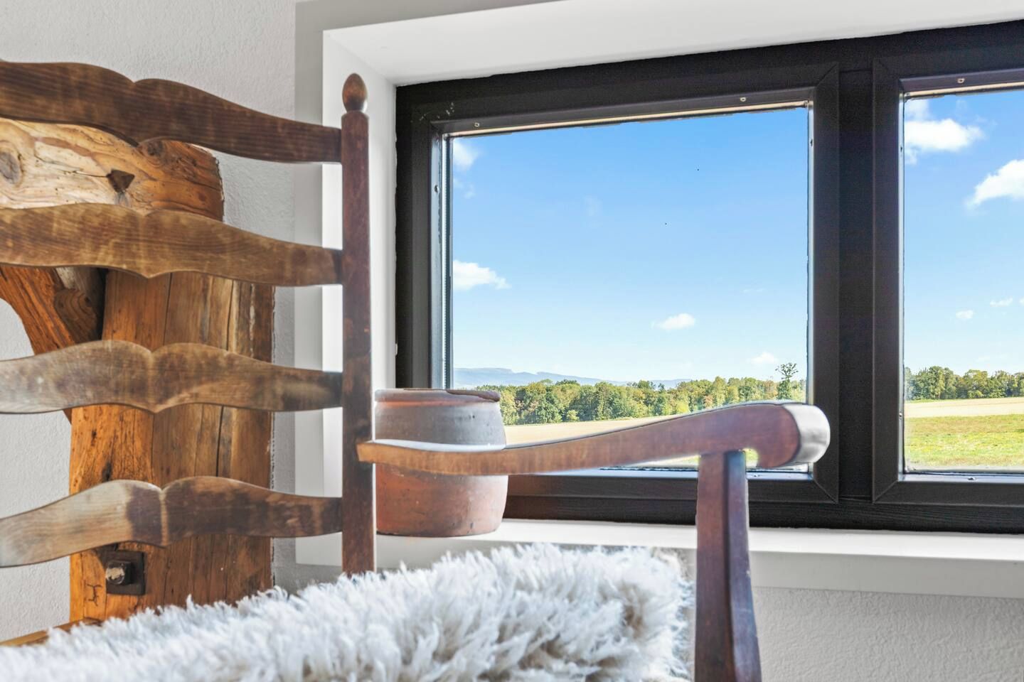 Ein hölzerner Stuhl mit rustikalem Design, ein weißer, flauschiger Teppich und ein Fenster mit Blick auf eine grüne Landschaft.