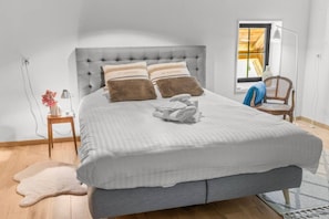 A neatly made bed with two pillows, a bedside table with a lamp, a chair, and a window with a view.