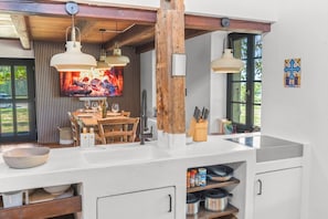 A modern kitchen with a wooden ceiling, pendant lights, a dining area with a TV, and a view of the outdoors.