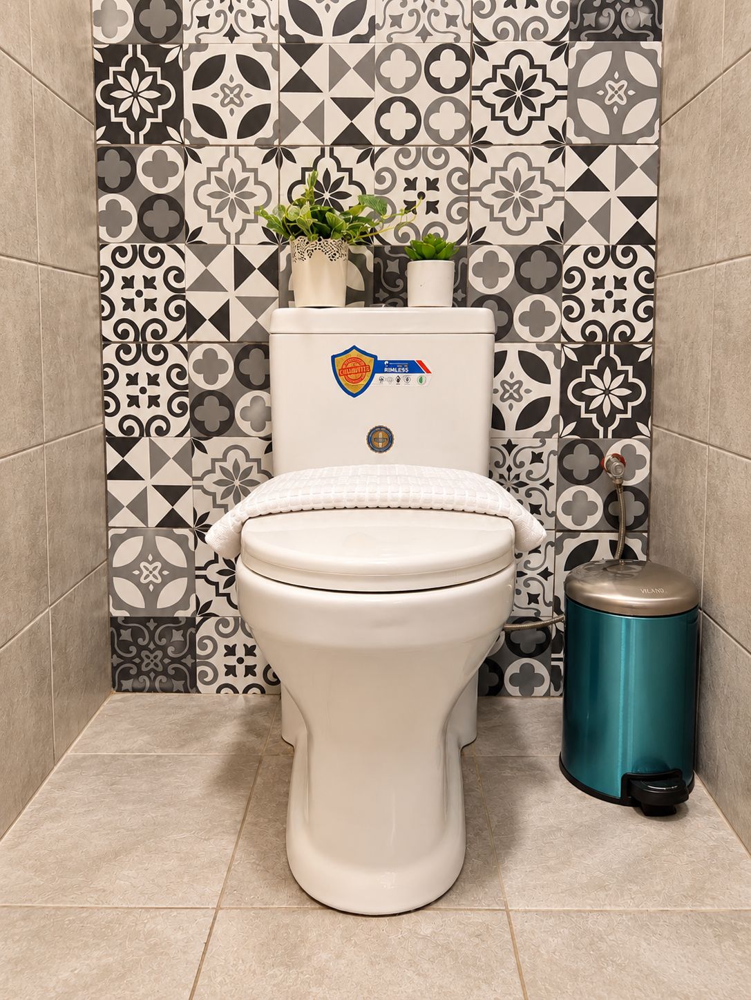 A toilet with a white seat and lid, a toilet paper holder, and a trash can in a bathroom with patterned tiles.