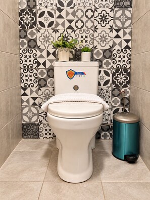 A toilet with a white seat and lid, a toilet paper holder, and a trash can in a bathroom with patterned tiles.