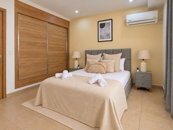 A bedroom with a bed, two bedside tables, an air conditioning unit, a framed picture on the wall, and a wooden wardrobe.