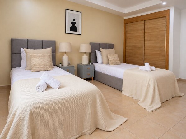A hotel room with two beds, each with a grey headboard, white bedding, and a beige throw blanket. There are two bedside tables with lamps and a framed picture on the wall.