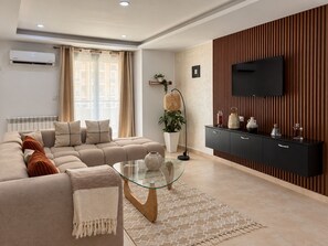Una sala de estar moderna con un sofá seccional, una mesa de centro de vidrio, un televisor de pantalla plana montado en una pared con paneles de madera y un radiador en la pared.
