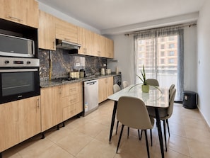 Una cocina moderna con armarios de madera, horno empotrado, lavavajillas y un comedor con mesa y sillas de vidrio.