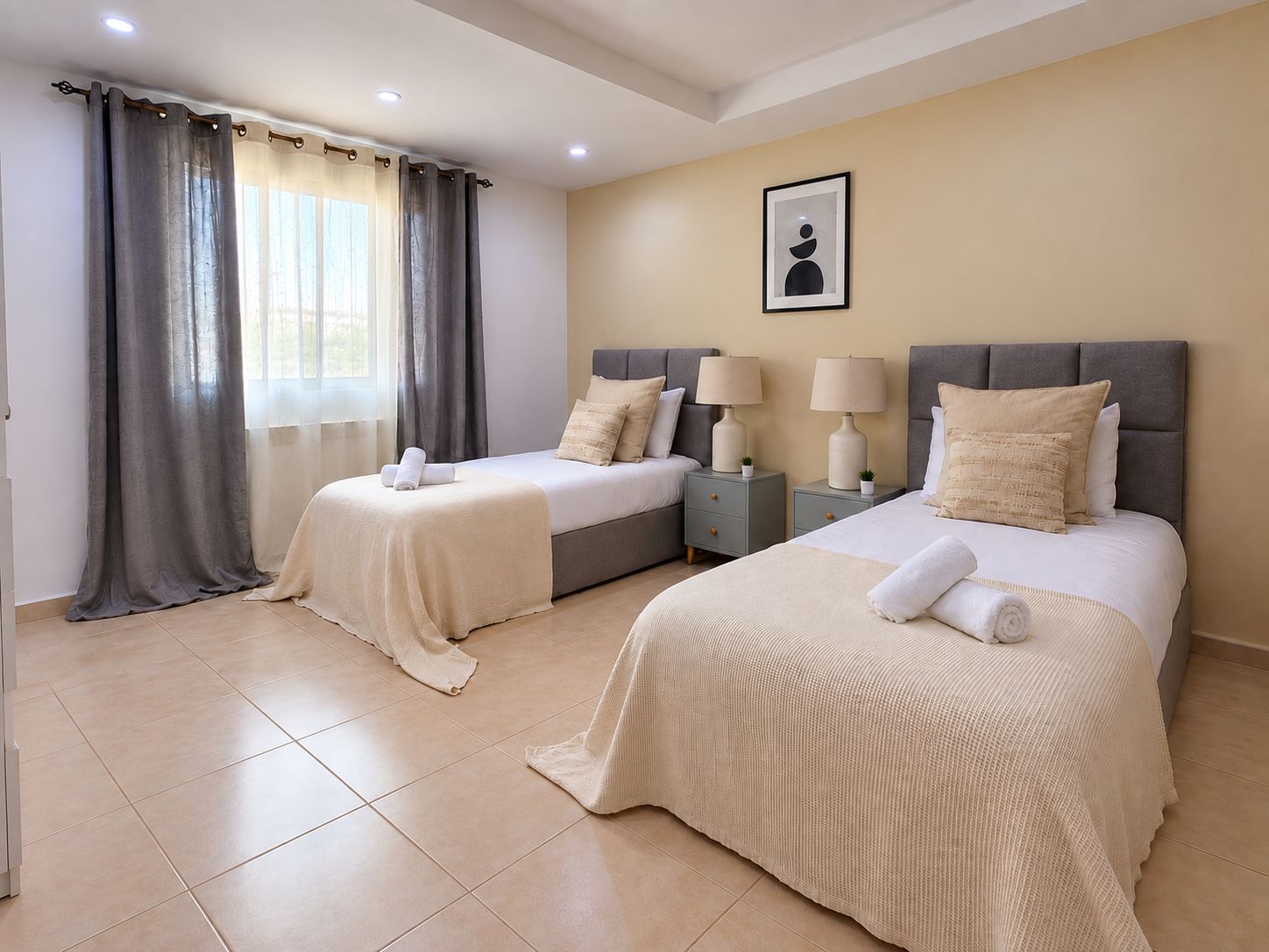 A hotel room with two beds, a large window with curtains, a framed picture on the wall, and a tiled floor.
