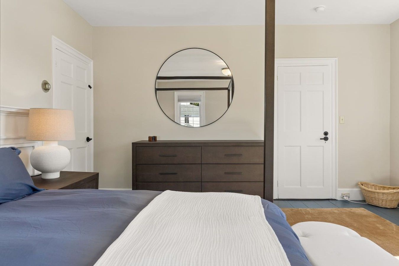 Een slaapkamer met een bed, een dressoir, een spiegel, een lamp en een deur.