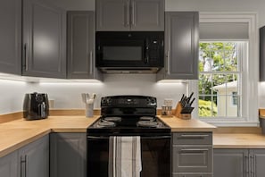 A kitchen with grey cabinets, a black microwave, a stove, and a window with a view of greenery.