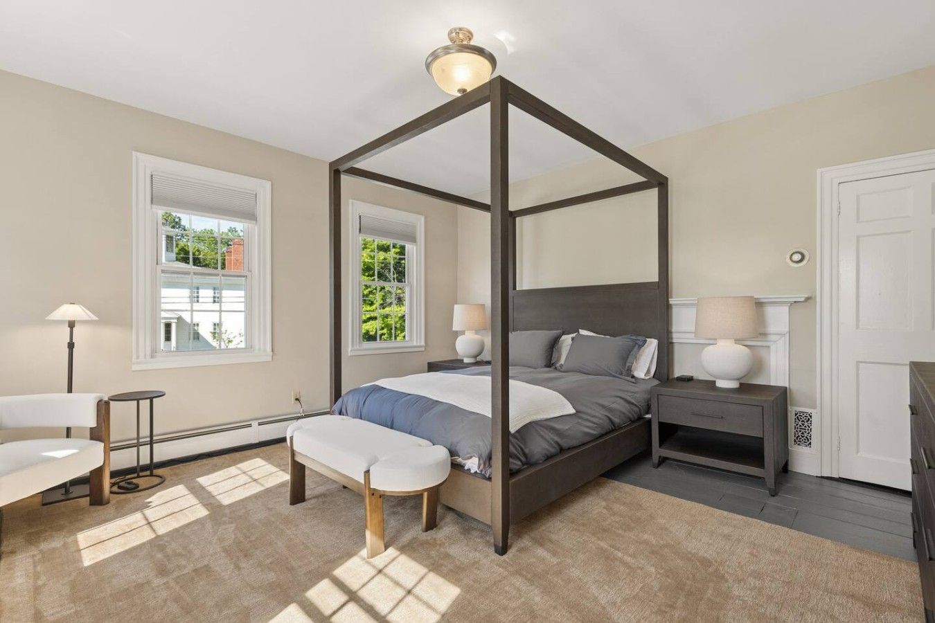 A four-poster bed with a nightstand and lamp, a bench, and a chair in a bedroom with large windows and a view of greenery.