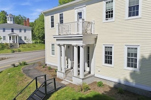 A two-story house with a front porch, a small staircase, and a road in the background. - The Federal Flat Steps From Downtown (Belfast)