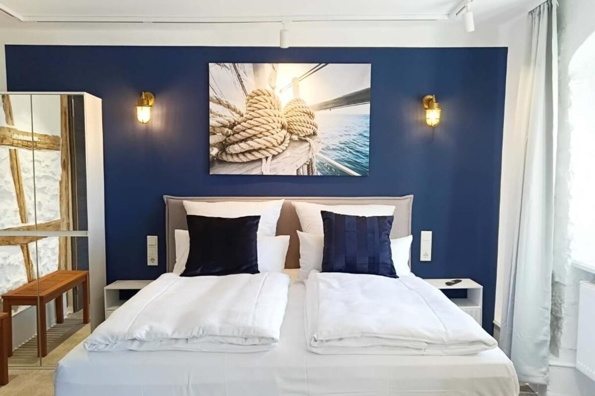 Una cama doble con ropa blanca, un cuadro abstracto grande encima y dos luces fijadas a la pared.
