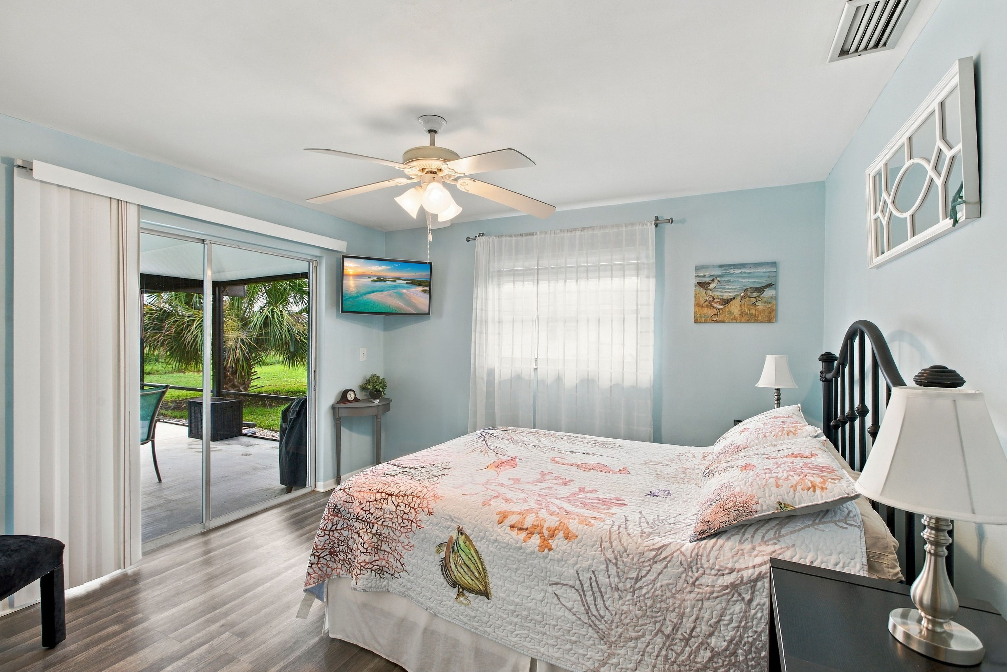 A bedroom with a bed, a ceiling fan, a window with curtains, a painting, and a view of the outdoors.