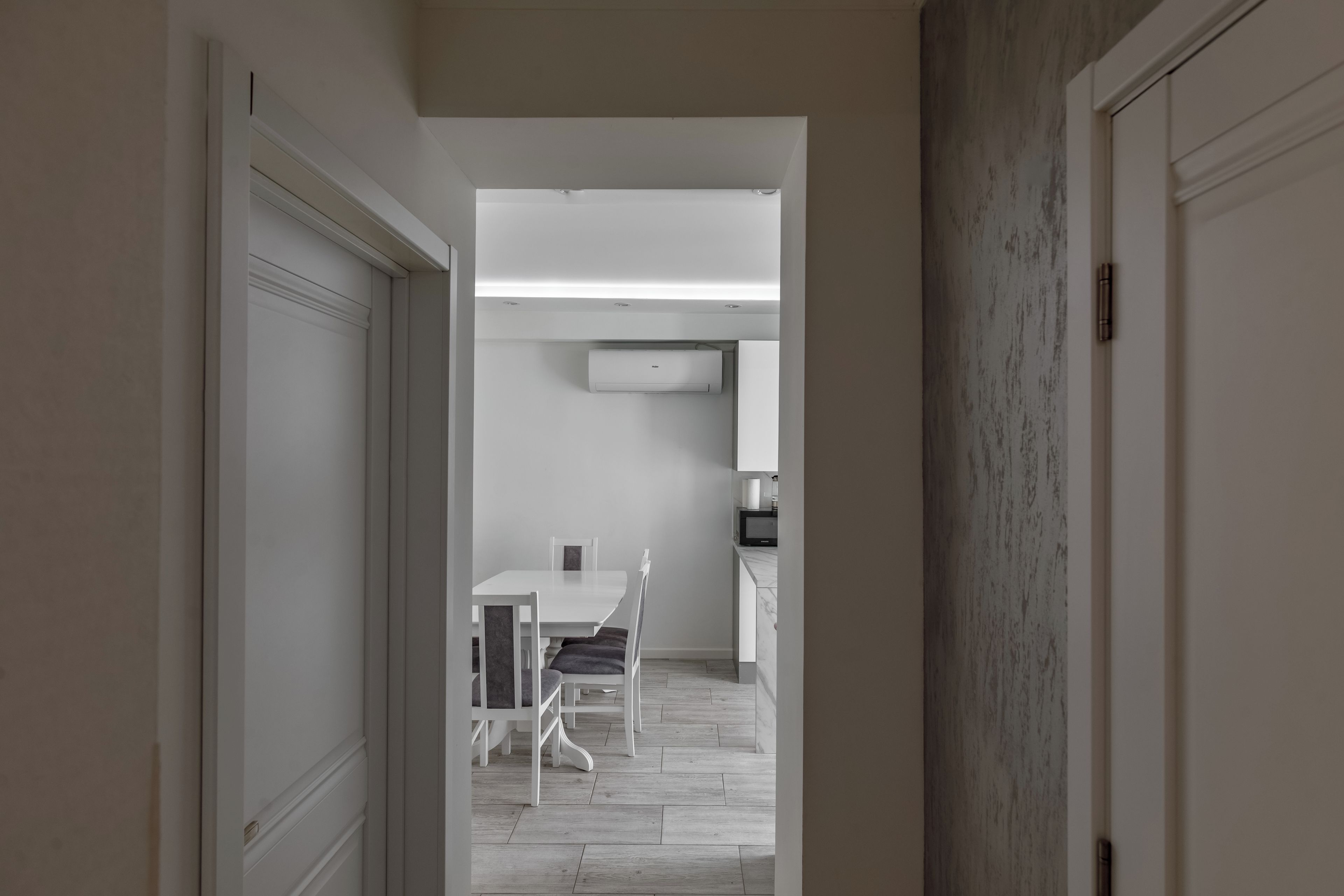 Un couloir mène à une salle à manger avec une table et des chaises blanches, une cuisine équipée d’un four à micro-ondes et un appareil de climatisation fixé au plafond.
