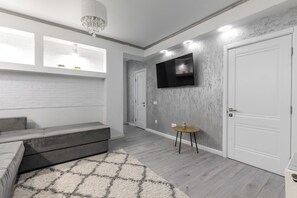 A modern living room with a grey sofa, a patterned rug, a flat-screen TV mounted on the wall, and a small round table with a vase.