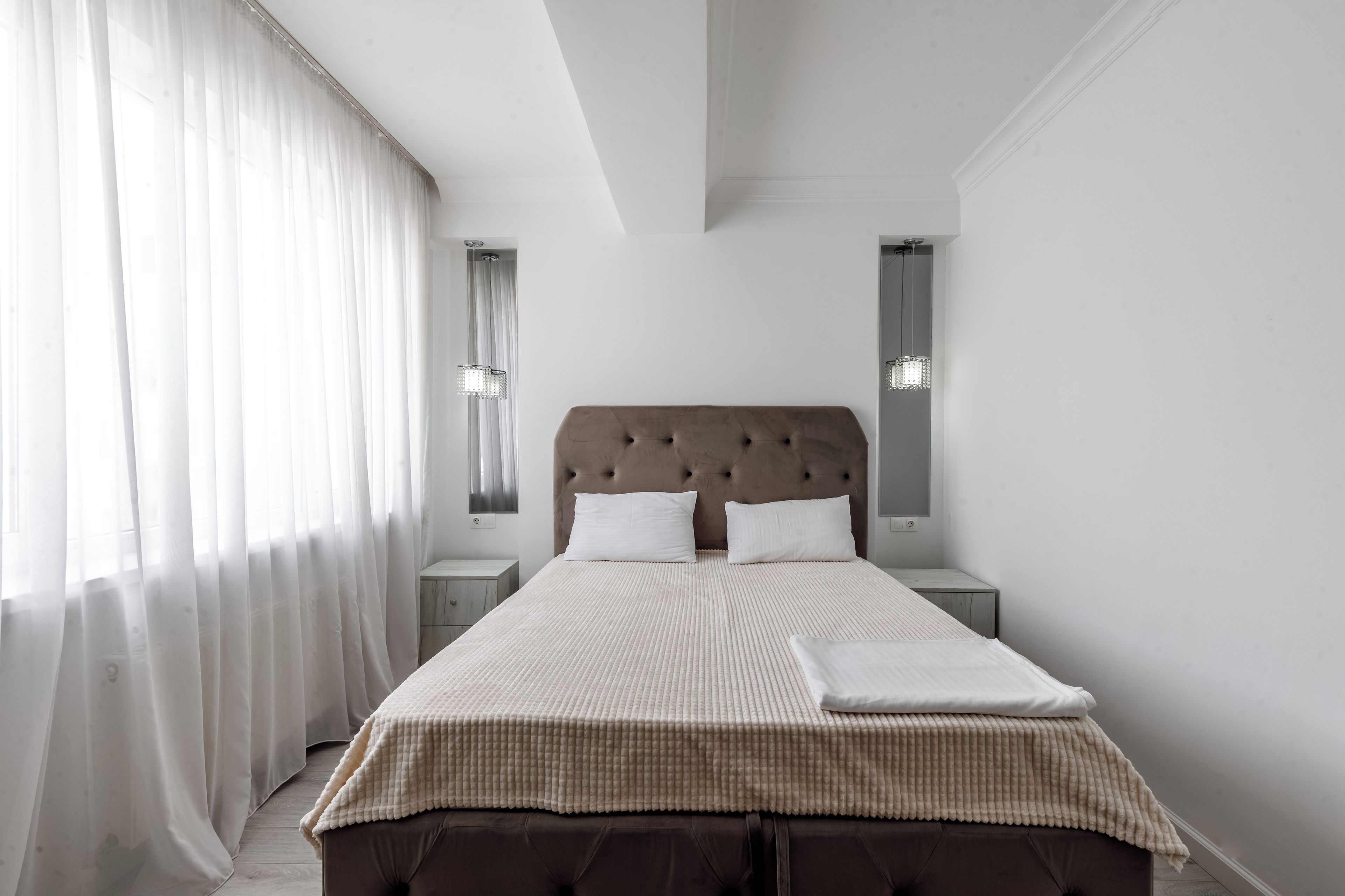 A bedroom with a large bed, a nightstand, a wall-mounted lamp, and sheer curtains.