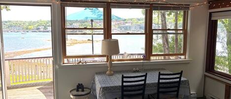 Una habitación con una mesa y sillas, ventanas amplias con vista a un cuerpo de agua y un muelle, y una mesita con una lámpara.
