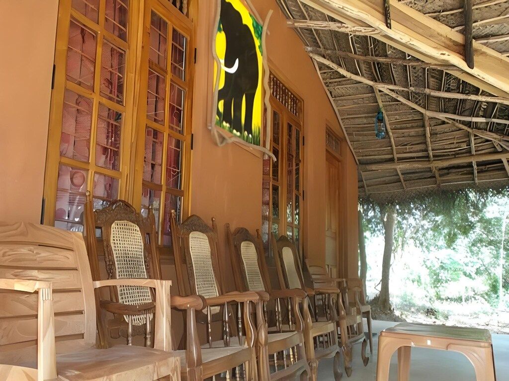 Un área de estar al aire libre techada con sillas de madera y una mesa, una ventana con un diseño de elefante en vitral y una estructura de techo de paja.