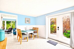 Een lichte kamer met een blauwe wand, een eettafel met stoelen, een raam met uitzicht op groen en een deur naar buiten.