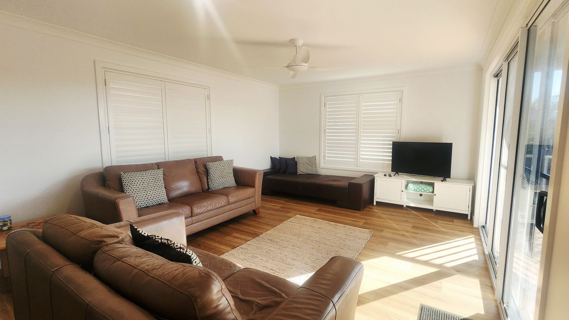 Un salon avec un canapé en cuir marron, un téléviseur posé sur un meuble blanc et un ventilateur de plafond.