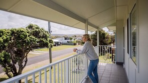 Un balcone con ringhiera, una persona che tiene in mano una tazza e vista su una strada e delle case.