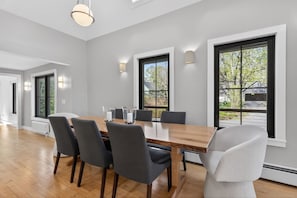 A modern dining room with a wooden table, grey chairs, and large windows offering a view of the outdoors.