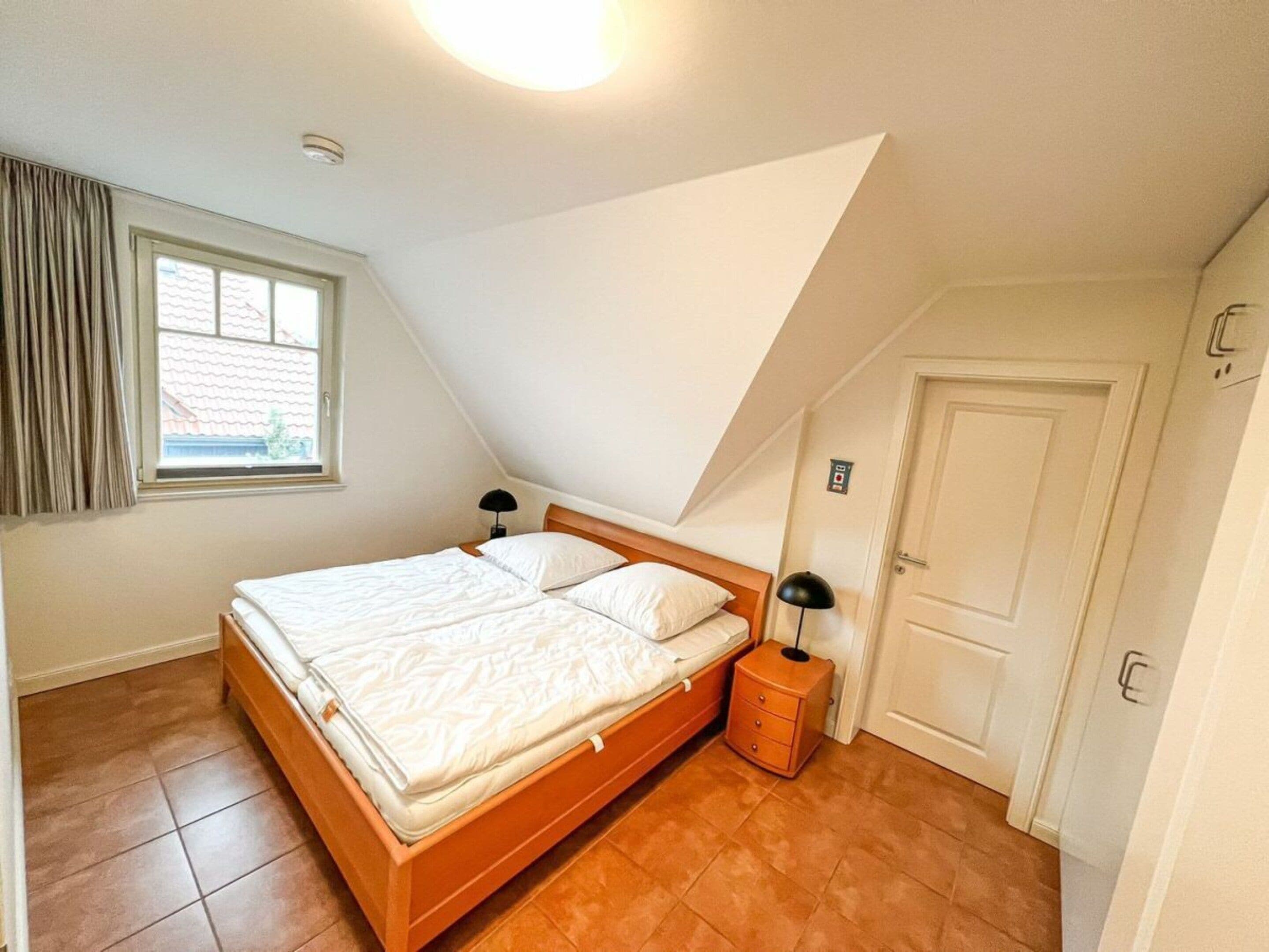 A bedroom with a sloped ceiling, a bed with white bedding, a bedside table with a lamp, and a window with curtains.