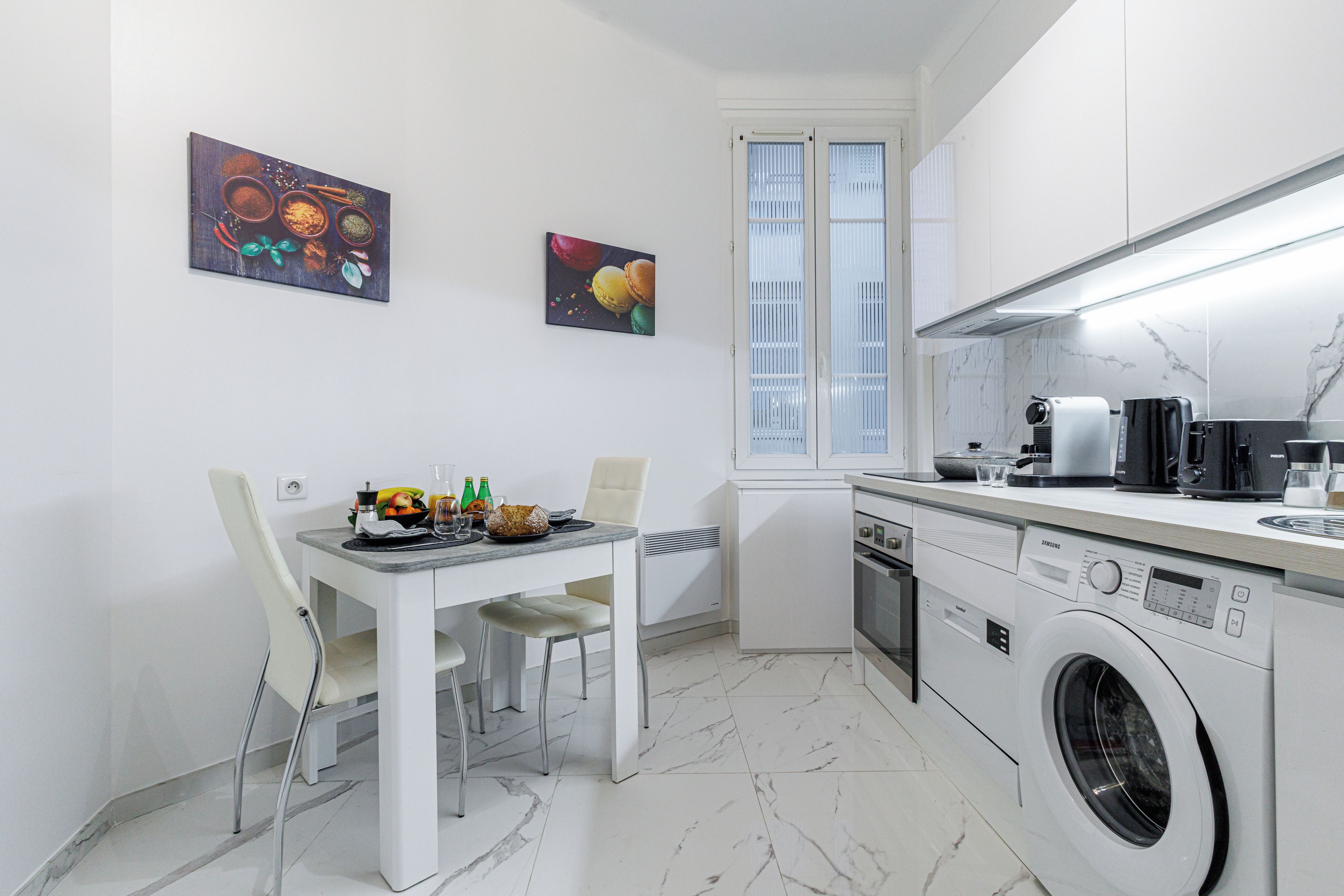 Une cuisine moderne avec un coin repas, des meubles blancs et un lave-linge, ainsi que deux œuvres d’art accrochées au mur.