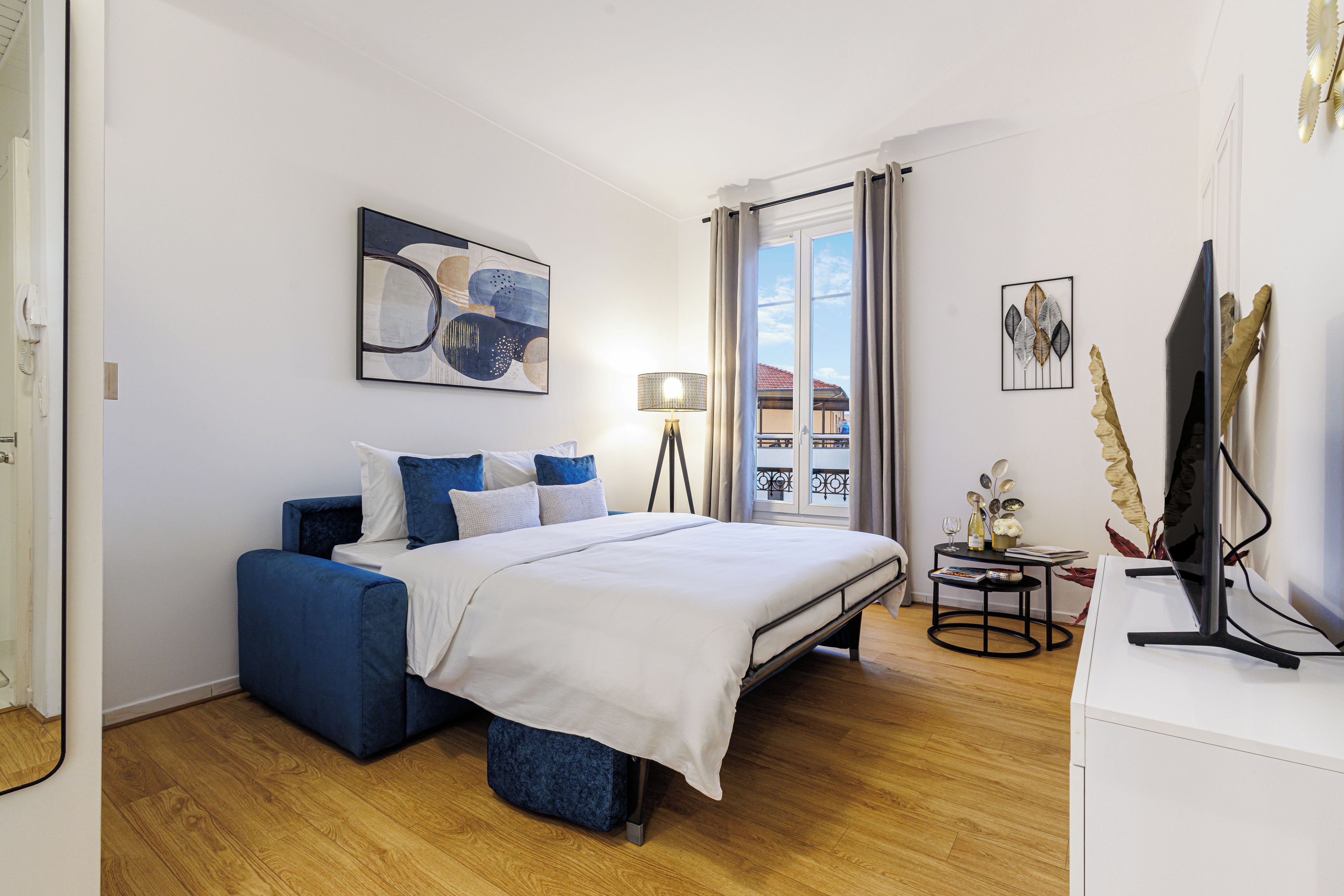 Ein modernes Schlafzimmer mit einem Bett, einem blauen Sofa, einem Schreibtisch mit Computer und Blick auf einen Balkon.