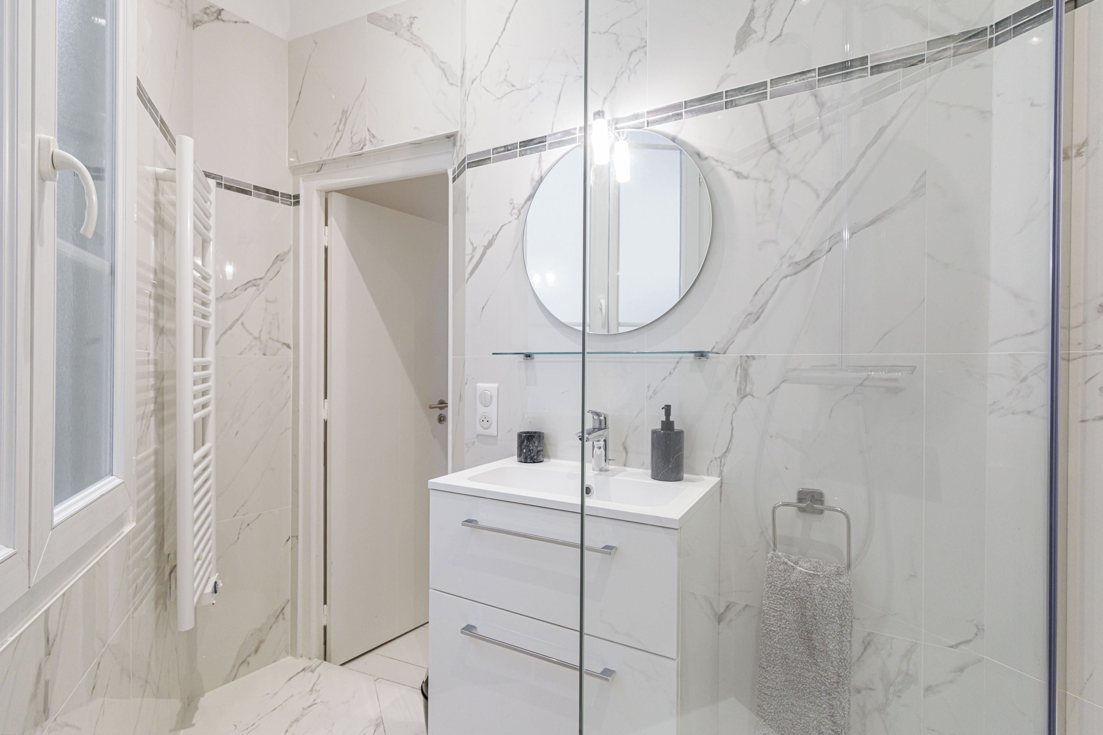 Une salle de bain moderne avec une douche à l’italienne en verre, un meuble-lavabo blanc avec un miroir rond et un mur recouvert de carreaux de marbre.