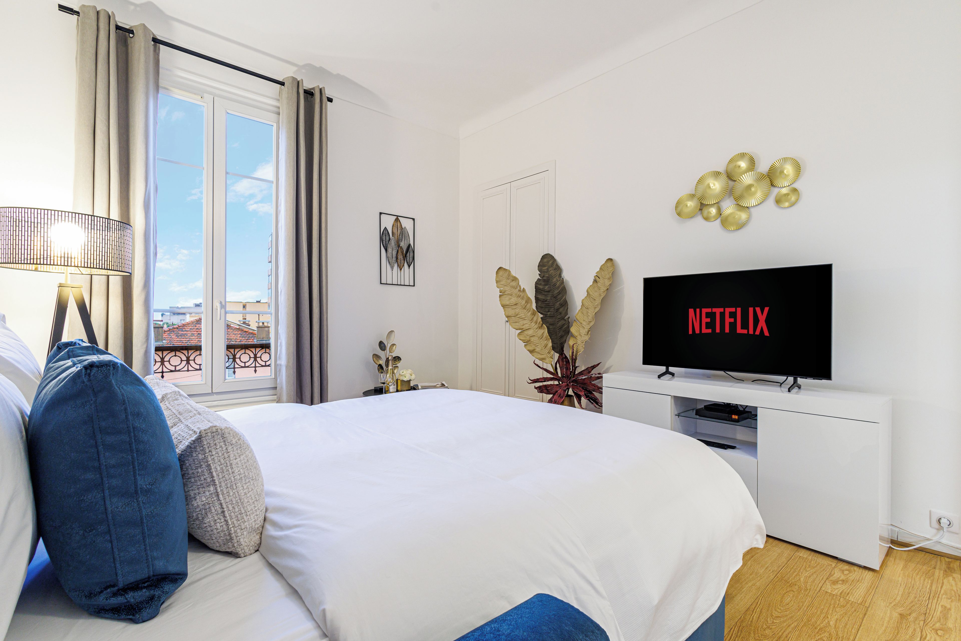 A hotel room with a bed, a TV displaying 'NETFLIX', a window with curtains, and a view of buildings outside.