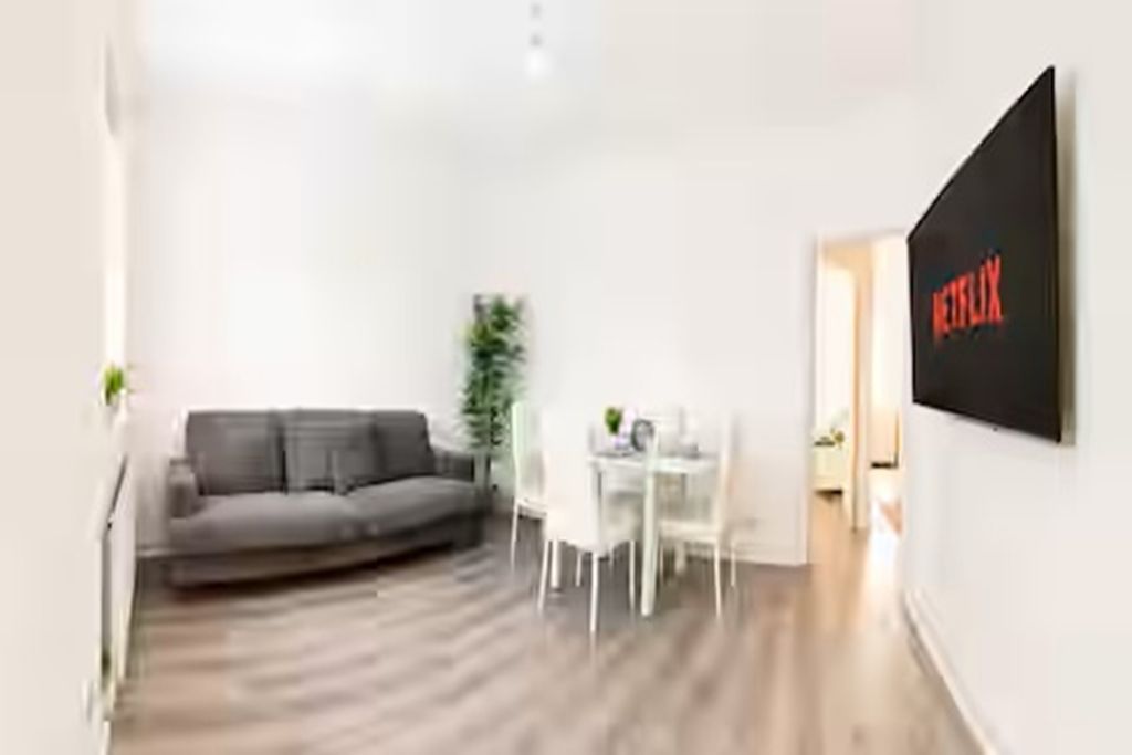 A modern living room with a grey sofa, a dining table with chairs, and a wall-mounted TV displaying the Netflix logo.
