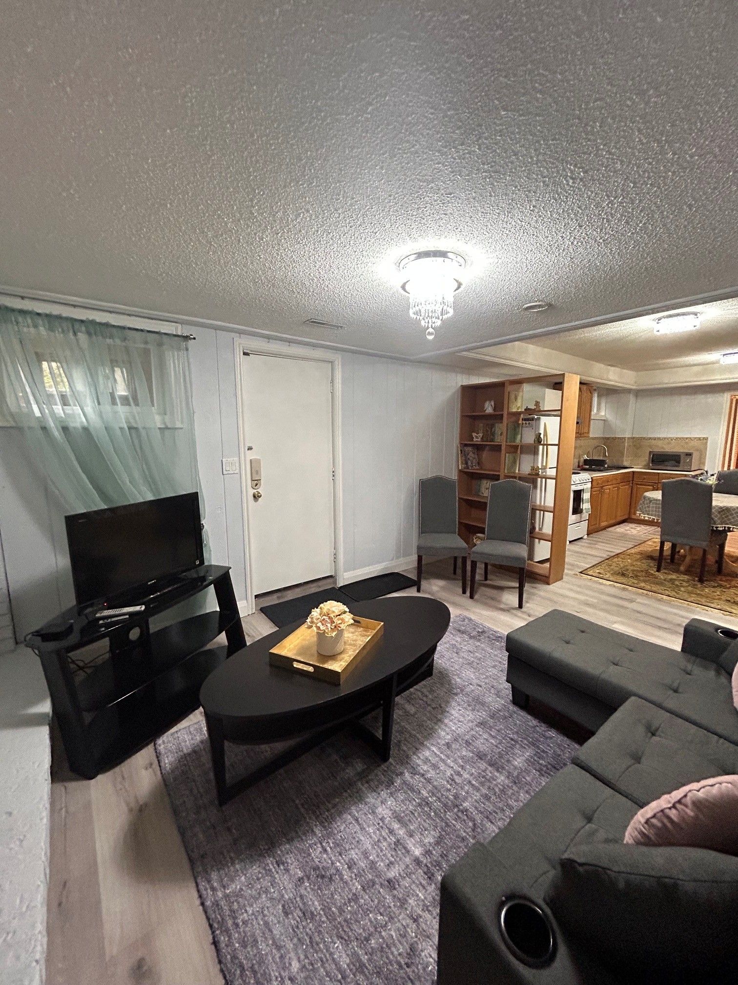 A living room with a TV, sofa, and coffee table. There is a kitchen in the background with a microwave and cabinets.