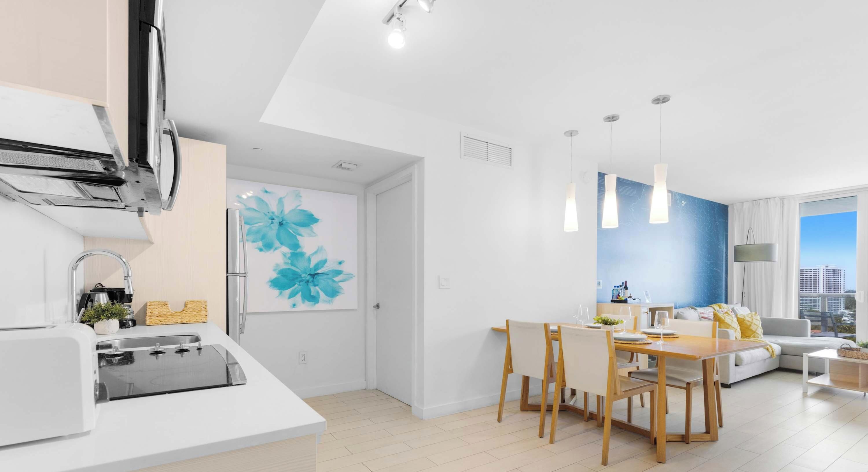 A modern kitchen with a built-in oven, a dining area with a wooden table and chairs, and a living room with a sofa and a large window.