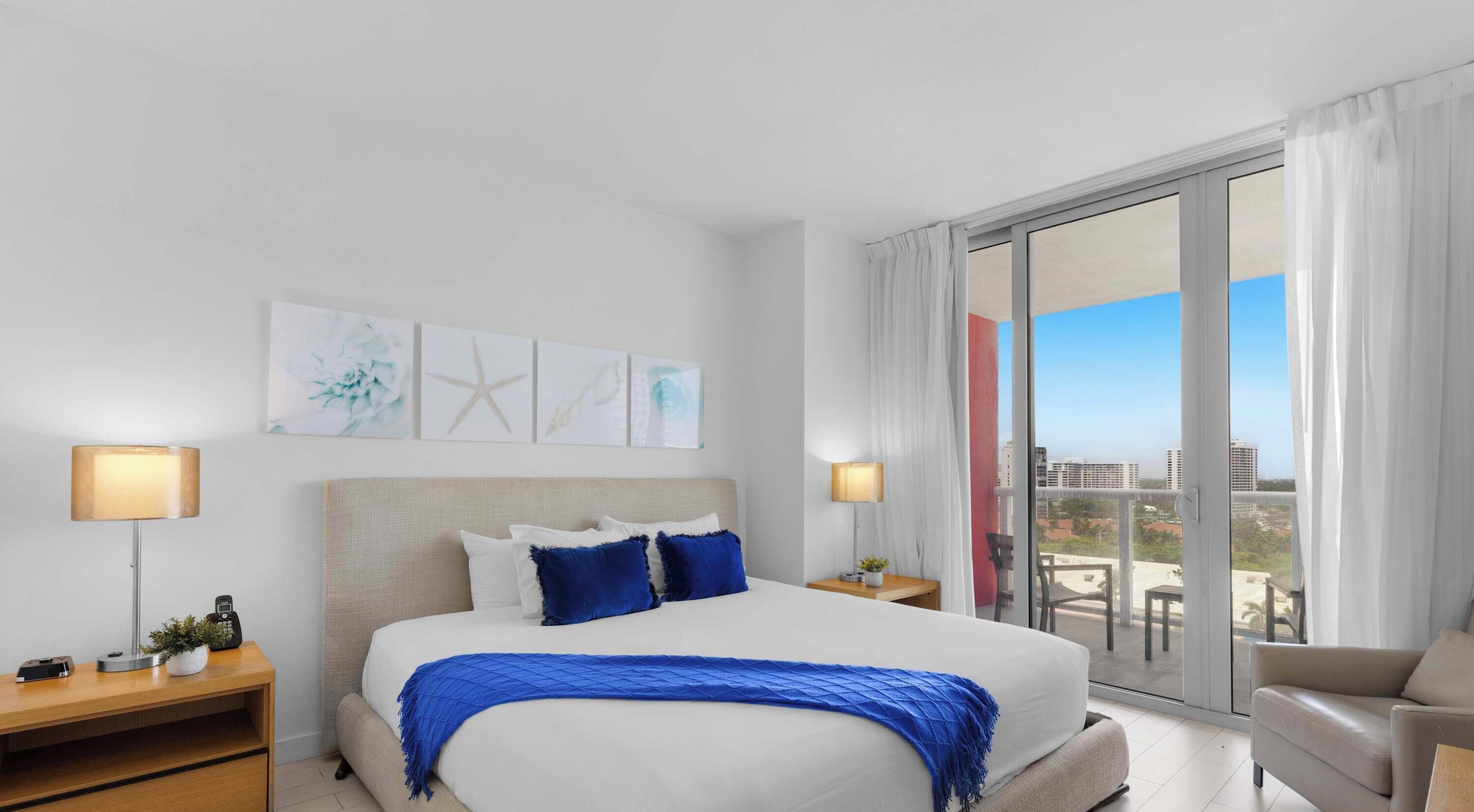Ein Hotelzimmer mit einem großen Bett, zwei Nachttischen, einem Sessel und einem Balkon mit Blick auf Gebäude.