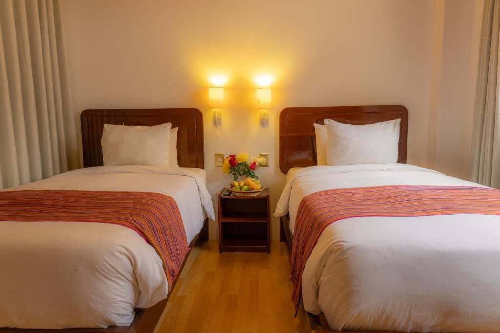 A hotel room with two beds, a nightstand with a vase of flowers, and wall-mounted lights.
