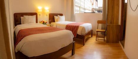 A hotel room with two beds, a wooden floor, a chair, and a window with curtains.
