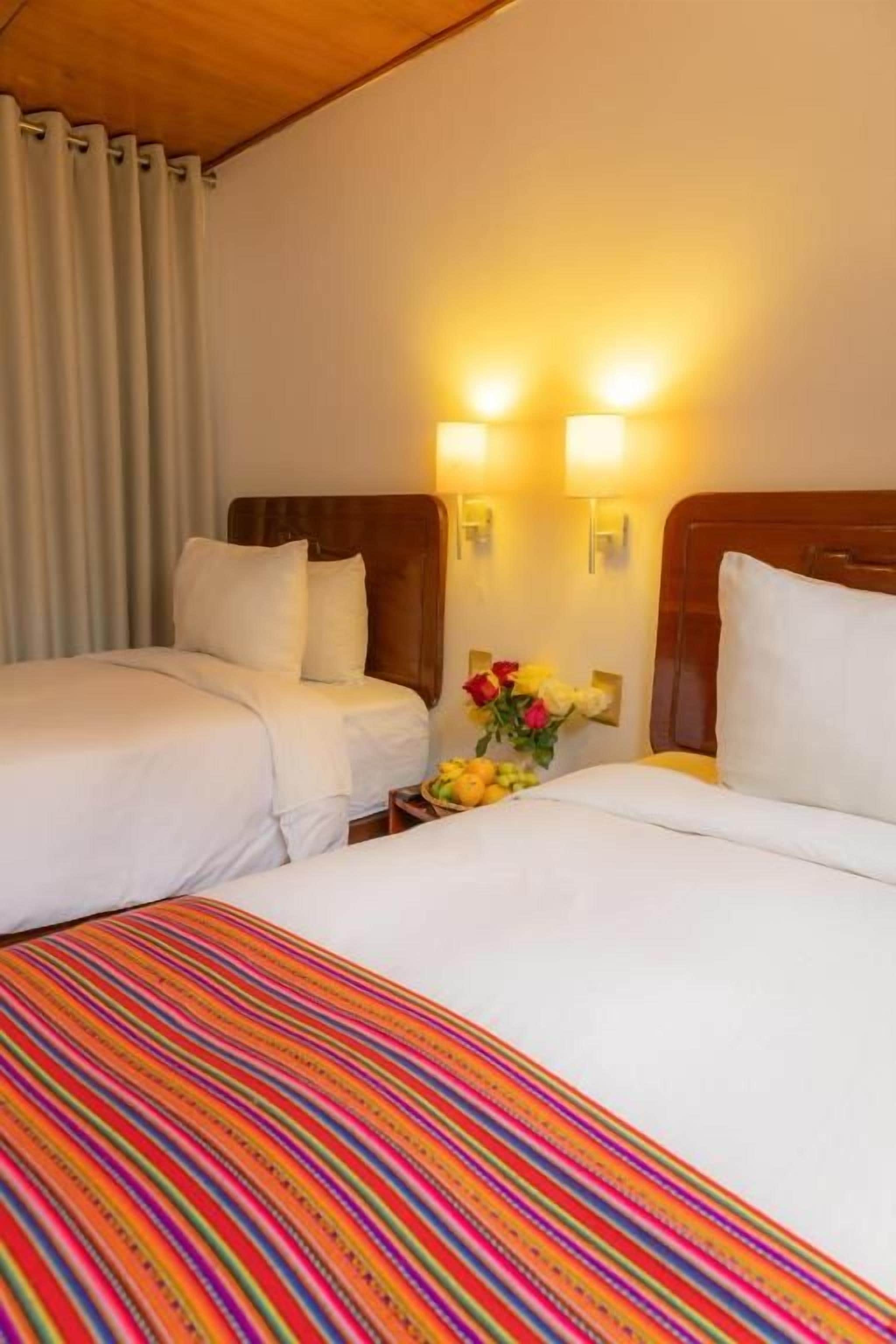 A hotel room with two beds, a wooden headboard, a nightstand with a vase of flowers, and wall-mounted lamps.