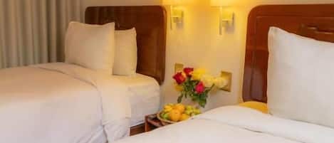 A hotel room with two beds, a wooden headboard, a nightstand with a vase of flowers, and wall-mounted lamps.