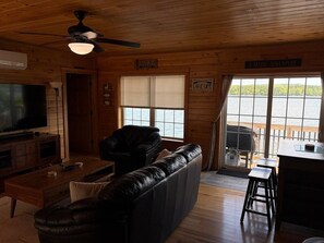 A cabin interior with a television, leather sofa, wooden furniture, and a view of the lake.