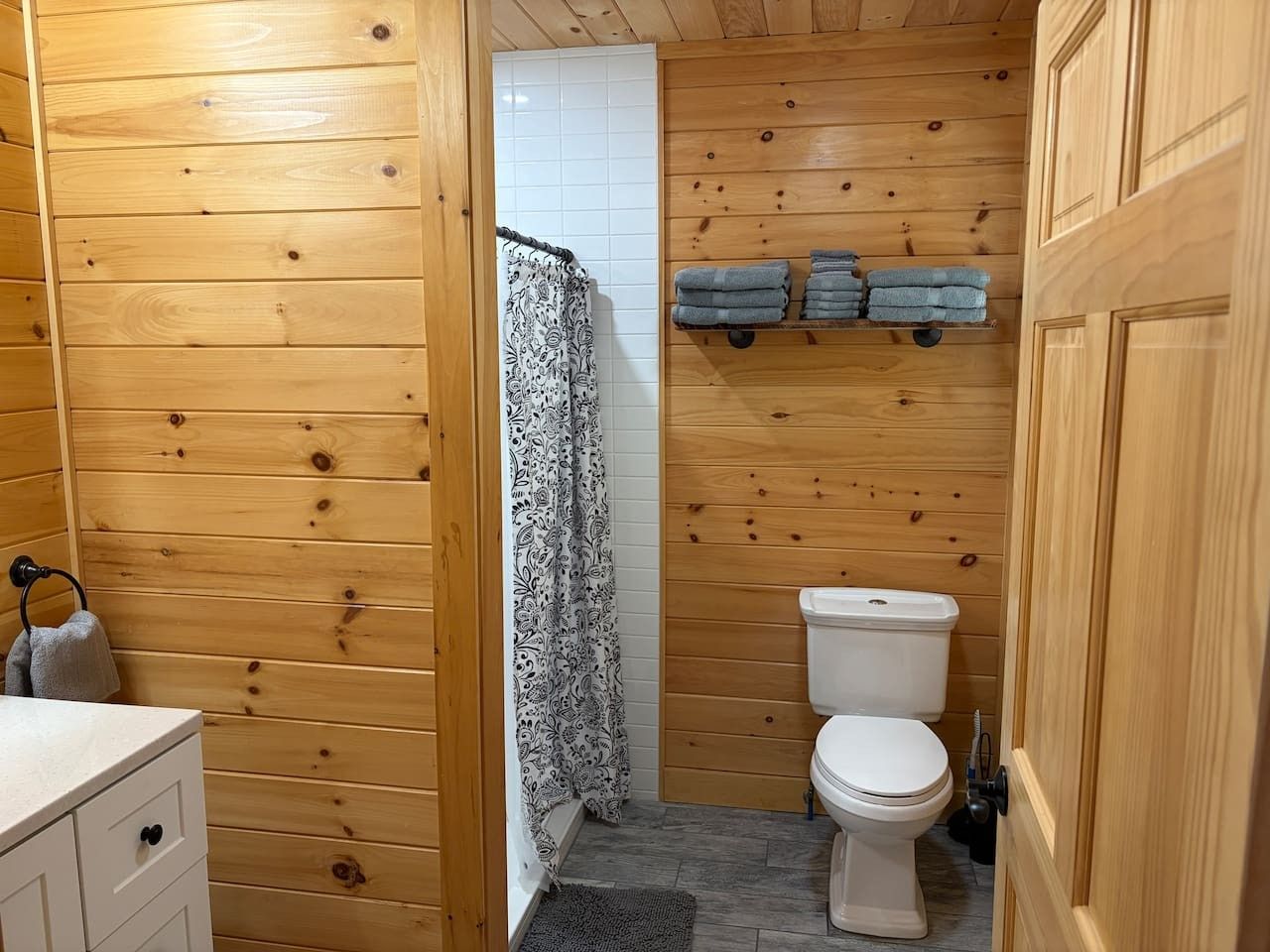 A wooden-paneled bathroom with a toilet, a towel rack, and a shelf with folded towels.