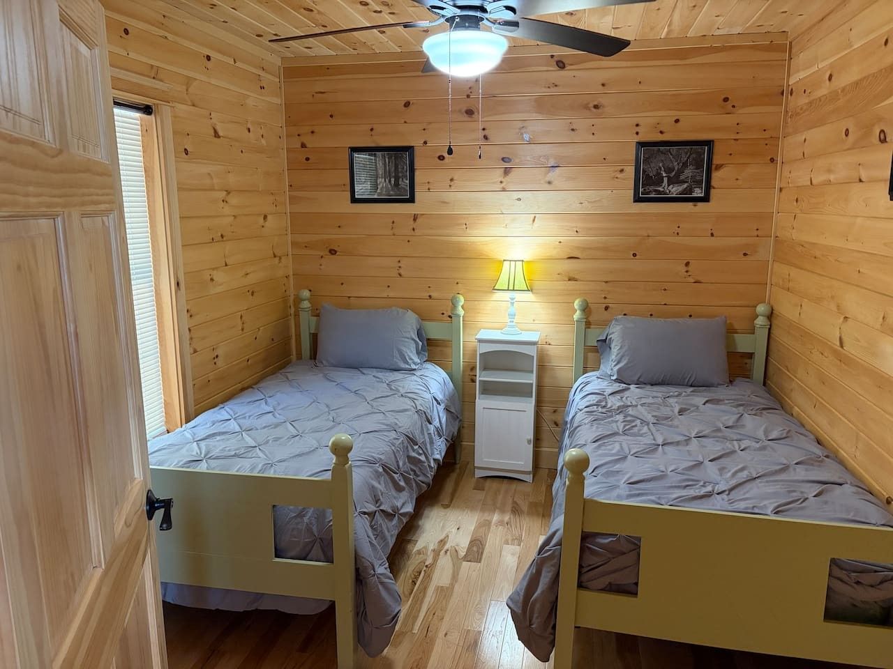 Quarto com duas camas de madeira, um ventilador de teto e uma mesa de cabeceira com luminária.