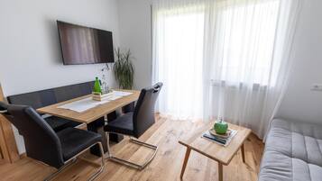 Ein modernes Wohnzimmer mit einem Holzes Esstisch, schwarzen Stühlen, einem grauen Sofa, einem an der Wand montierten Fernseher und einem großen Fenster mit durchsichtigen Vorhängen.