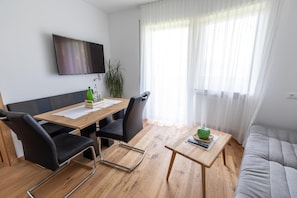 Ein modernes Wohnzimmer mit einem Holzes Esstisch, schwarzen Stühlen, einem grauen Sofa, einem an der Wand montierten Fernseher und einem großen Fenster mit durchsichtigen Vorhängen.