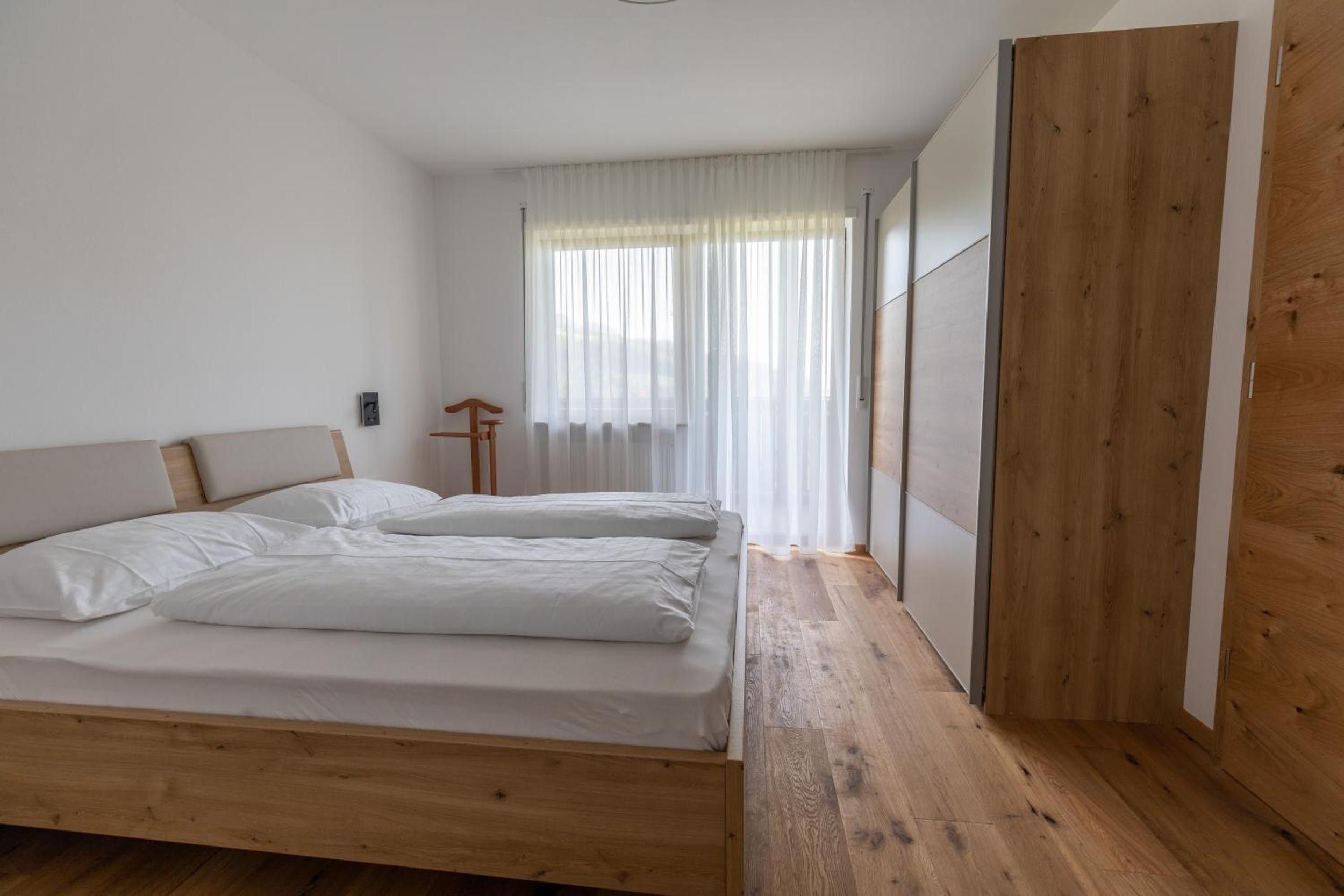 A bedroom with two beds, wooden flooring, a large window with sheer curtains, and a wooden panel on the wall.