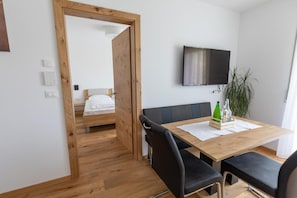 Ein Zimmer mit Holztür, einem Bett, einem an der Wand befestigten Fernseher, einem Esstisch mit Stühlen und einer Topfpflanze.