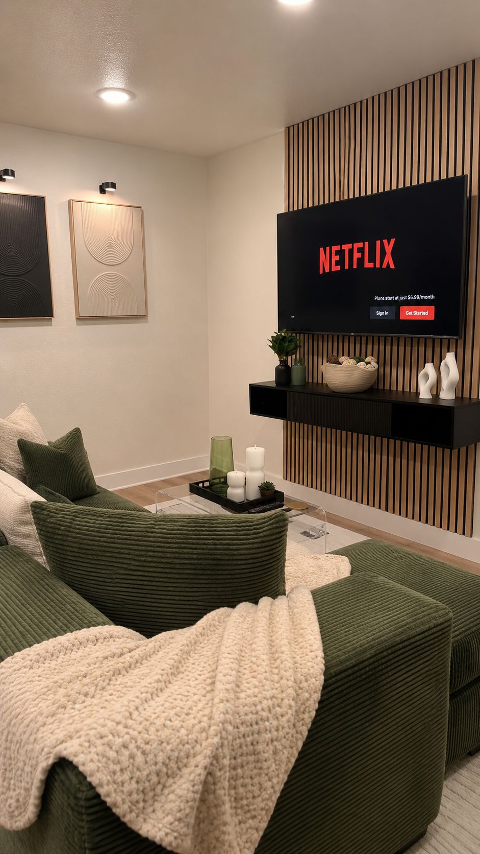 Un salon moderne avec un grand téléviseur à écran plat fixé au mur, un ensemble de canapés, des œuvres d’art décoratives et une petite table ornée de bougies et d’une plante.