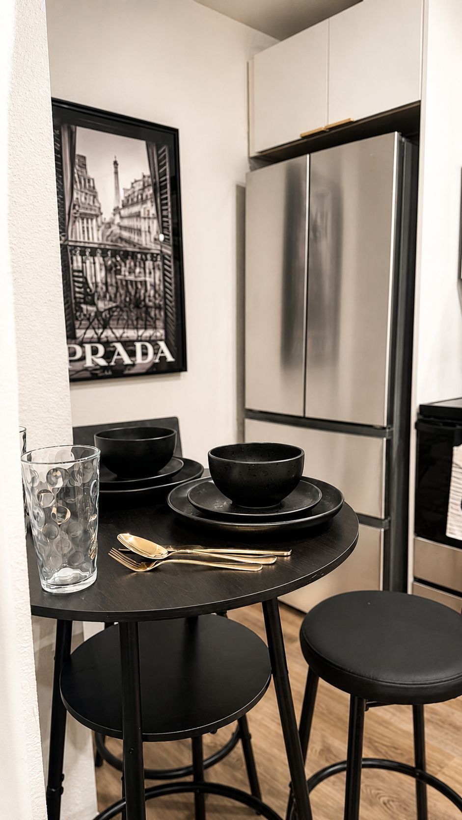 Een kleine keuken met een roestvrijstalen koelkast, een zwarte tafel met een kom en bestek, en een lijst met een foto aan de muur.