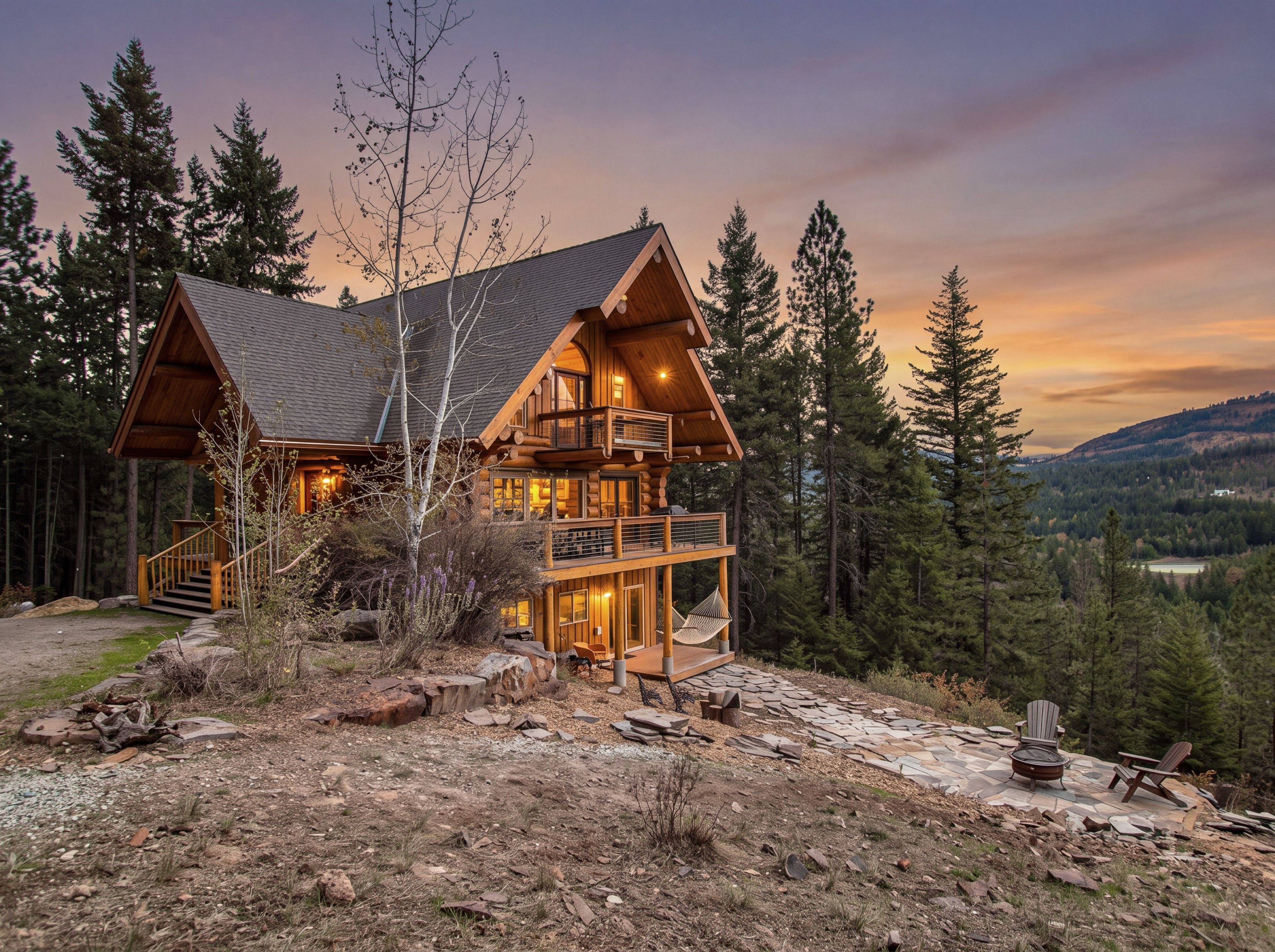 A log cabin with a balcony, surrounded by trees and a stone patio with outdoor furniture.