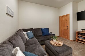 Een woonkamer met een hoekbank, een klein houten bijzettafeltje, een aan de muur bevestigde televisie en een deur.